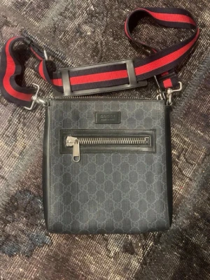 Svart axelväska - Snygg svart axelväska  med klassiskt GG-mönster och en röd- och blårandig justerbar axelrem. Väskan har en dragkedjeficka framtill och detaljer i skinn. Perfekt för att bära det viktigaste med stil.
