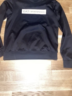 Svart sweatshirt från EA7 Emporio Armani - Snygg svart sweatshirt från EA7 Emporio Armani med vit logga på bröstet. Tröjan har rund halsringning, långa ärmar och ribbade muddar vid ärmslut och nederkant. Perfekt för en stilren och avslappnad look.