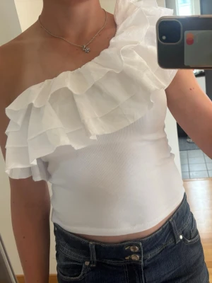 Vit oneshoulder topp med volang - Snygg vit oneshoulder topp med bred volangdetalj över axeln. Toppen är ärmlös och har en tight passform som framhäver figuren. Perfekt för dig som vill sticka ut med en trendig och unik look. Toppen har fått en liten missfärgning vid armvecket på ena sidan, men den är knappt använd. 