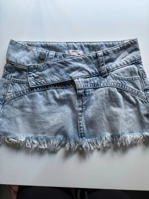 Ljusblå jeanskjol från Gina Tricot - Trendig ljusblå jeanskjol från Gina Tricot i kort modell. Kjolen har fransig nederkant, sned knappdetalj och klassiska fickor fram. Perfekt för dig som gillar en avslappnad och cool stil.
