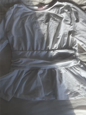 Vit långärmad klänning Gina Tricot - Vit topp från Gina Tricot med lång ärm, rund hals och markerad midja. Kjolen har volang och modellen är enkel men stilren. Tillverkad i mjuk bomull som känns skön mot huden.