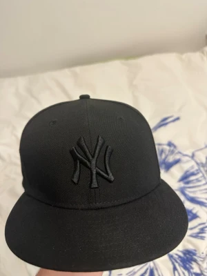 Svart New York Yankees keps - Snygg svart keps från New Era med New York Yankees-logga broderad framtill i svart. Klassisk rak skärm och broderad MLB-logga baktill. Tillverkad i ett tåligt tyg som passar perfekt till streetwear och casual outfits. Storlek:7 3/8 =58,7 cm