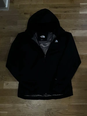 Svart regnjacka från The North Face - Snygg svart regnjacka från The North Face med huva och dragkedja framtill. Jackan har meshfoder på insidan och logga på bröstet. Perfekt för regniga dagar och enkel att matcha med olika outfits.