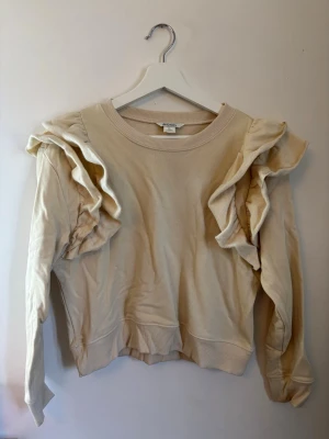 Beige sweatshirt med volangärmar - Säljer en beige sweatshirt med unika volangdetaljer längs båda ärmarna. Tröjan har rund halsringning, ribbade muddar och ribbad underkant. Perfekt för dig som vill ha en basic tröja med en twist.