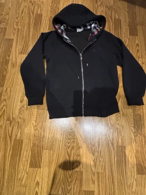 Svart hoodie med dragkedja och huva - Säljer en svart hoodie med dragkedja och huva. Insidan av huvan har ett rutigt mönster i svart, vitt och rött. På bröstet finns en diskret broderad logga. Hoodien har långa ärmar och ribbade muddar. Perfekt för en chill och stilren look.