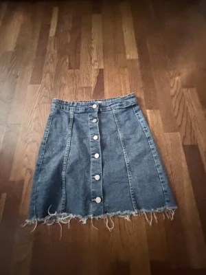 Kort jeanskjol med knappar framtill - Säljer en blå jeanskjol med rå kant och knappar längs hela framsidan. Kjolen har en A-linjeformad passform och tydliga sömmar som ger en snygg vintagekänsla. Perfekt till sneakers eller boots för en avslappnad look.
