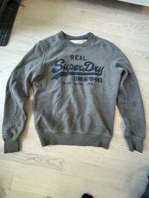 Grå sweatshirt från Superdry - Snygg grå sweatshirt från Superdry med mörkblå logga och japansk text på bröstet. Tröjan har rund halsringning, långa ärmar och ribbade muddar vid ärmslut och nederkant. Perfekt för en avslappnad och trendig stil.