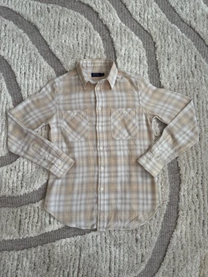 Beige och vit rutig skjorta - Snygg rutig skjorta i beige och vitt med två bröstfickor och klassisk krage. Skjortan har långa ärmar och knäppning framtill. Perfekt för en avslappnad men stilren look.