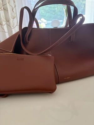 Brun handväska i läder  - Snygg brun handväska  i slätt läder med matchande clutch. Väskan har dubbla handtag, vita kontrastsömmar och minimalistisk design. Clutchen har guldfärgad dragkedja och präglad logga. Perfekt för dig som gillar stilrena accessoarer.