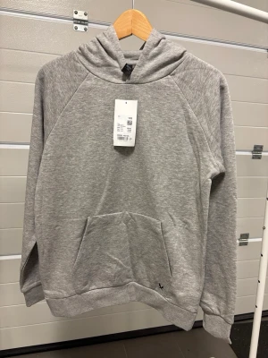 Ljusgrå tunn hoodie med huva - En stilren tunn ljusgrå hoodie med huva och känguruficka framtill. Tillverkad i mjukt sweatshirtmaterial och har långa ärmar samt ribbade muddar vid ärmslut och nederkant. Perfekt för en avslappnad och enkel look.