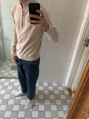 Half zip gant - Snygg beige stickad tröja med halv dragkedja och hög krage. Tröjan har ribbade muddar vid ärmslut och nederkant samt en liten broderad logga på bröstet. Perfekt för lager på lager och passar till jeans eller chinos.