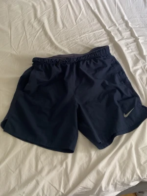 Mörkblå träningsshorts från Nike - Snygga mörkblå träningsshorts från Nike med elastisk midja och diskret logga på ena benet. Shortsen har en enkel design och är tillverkade i ett lätt syntetmaterial som passar perfekt för aktiva dagar. (Fickan där bak går it att stänga)