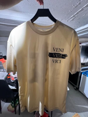 Beige t-shirt med VENI VIDI VICI-tryck - Säljer en beige t-shirt med svart texttryck 'VENI VIDI VICI' på bröstet och ett stort konstnärligt motiv på ryggen. T-shirten har rund hals och korta ärmar. Perfekt för dig som gillar unika prints och stilrena färger. Finns att hämta i Gröndal.