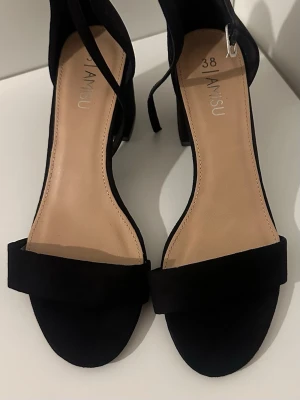 Svarta klackpumps  - Stilrena svarta pumps med öppen tå och bred rem över foten. Skorna har en klack och smala ankelband som knyts runt vristen. Materialet ser ut att vara mockaimitation och innersulan är beige. Perfekta för att lyfta din outfit.