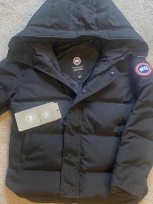 Canada Goose Macmillan - Snygg svart dunjacka från Canada Goose med klassisk patch på ärmen. Jackan har huva, dragkedja och knappar framtill samt två sidofickor. Perfekt för kalla vinterdagar och riktigt varm tack vare dunfyllningen.