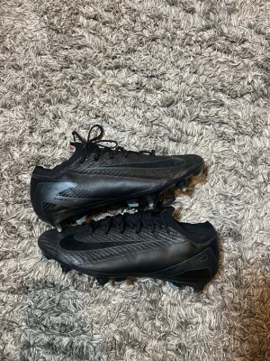 Svarta Nike fotbollsskor med dobbar - Säljer ett par svarta Nike fotbollsskor med låg profil och snörning. Skorna har en modern design med syntetiskt material och slitstarka dobbar för bra grepp på planen. Swoosh-logga på sidan och undersidan. Perfekta för dig som vill ha stilrena och snabba skor på matchen.