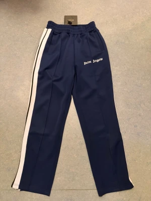 Blåa mjukisbyxor Palm Angels - Snygga blåa mjukisbyxor från Palm Angels med vita sidoränder och broderad logga på ena benet. Byxorna har elastisk midja och raka ben, perfekta för en chill och trendig look.  Storlek S/M alltså att den passar i S och M 