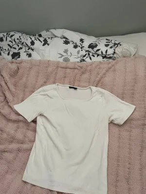 Vit ribbad t-shirt med fyrkantig ringning - En stilren vit t-shirt med ribbad struktur och fyrkantig ringning. T-shirten har korta ärmar och är tillverkad i ett mjukt material som känns skönt mot huden. Perfekt att matcha med jeans eller kjol för en enkel och clean look. Säljer för att den inte passar längre men är knappt använd och i bra skick.