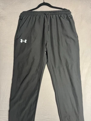 Svarta mjukisbyxor från Under Armour - Snygga svarta mjukisbyxor från Under Armour med elastisk midja och dragsko. Byxorna har en avslappnad passform och det klassiska Under Armour-loggan i vitt på vänster ben. Perfekta för chill eller träning.