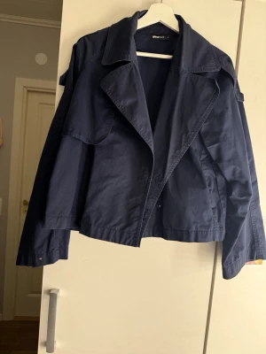 Mörkblå trenchcoat med bred krage - Snygg mörkblå trenchcoat med bred krage och knappar framtill. Jackan har en avslappnad passform och är tillverkad i ett tunt, vävt material som passar perfekt till vår och höst. Klassisk design med axelklaffar och långa ärmar.  Jackan har lite fläckar.
