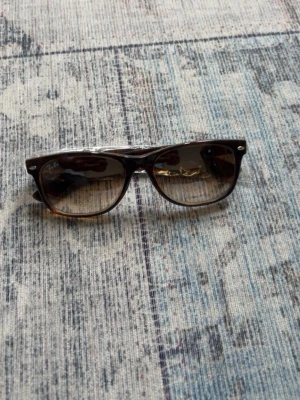  Ray-Ban solglasögon  - Snygga Wayfarer solglasögon från Ray-Ban i brunmelerad plast med mörka glas. Klassisk fyrkantig form och diskreta silvriga detaljer vid bågens framkant. Perfekta för att lyfta din stil under soliga dagar.