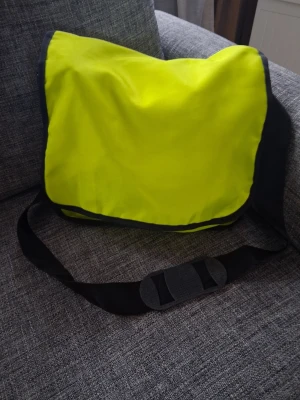 Neongul axelväska med svart kant - Axelväska i starkt neongult tyg med svarta kanter och justerbar axelrem. Väskan har en klassisk rektangulär form och stängs med ett lock. Perfekt för dig som vill sticka ut och bära dina grejer smidigt.