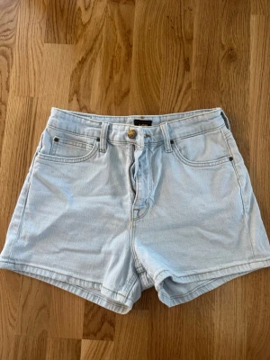 Ljusblå denimshorts från Lee - Snygga ljusblå denimshorts från Lee med klassisk femficksdesign och hög midja. Shortsen har bälteshällor, knappgylf och Lee-logga på bakfickan. Perfekta för sommaren och tillverkade i slitstarkt bomullsmaterial.