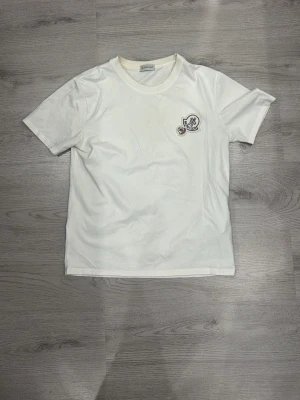 Vit t-shirt med Moncler-logga - Snygg vit t-shirt från Moncler med rund hals och korta ärmar. På bröstet sitter en broderad Moncler-logga i vitt, rött och blått. Tillverkad i mjukt bomullsmaterial som känns skönt mot huden. Perfekt till jeans eller shorts för en clean och stilren look.