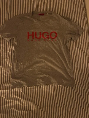 Grå Hugo Boss t-shirt med röd logga - Snygg grå t-shirt från Hugo Boss med stor röd HUGO-logga på bröstet. Klassisk rund hals och korta ärmar. Tillverkad i mjukt bomullsmaterial som känns skönt mot huden. Perfekt för en avslappnad och stilren look.