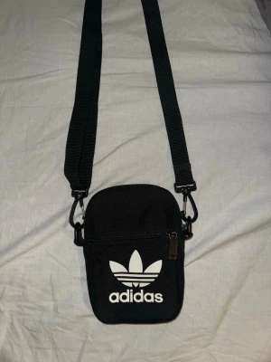 Adidas axelväska - Svart axelväska från Adidas med justerbar rem och klassisk vit trefoil-logga framtill. Väskan har dragkedja och är tillverkad i slitstarkt tygmaterial. Perfekt för att bära det viktigaste och har en stilren, sportig look.