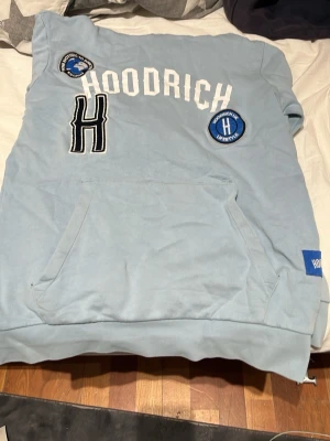 Hoodrich hoodie  - En skit snygg tröja från hoodrich, tröjan är ljusblå och har en snygg logga på bröstet. Tröjan är användbar jag själv har använt den några gånger, vid fickorna på magen har ena sömnen gått sönder men det är sytt syns om man kollar noga.