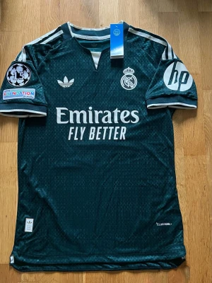 Real Madrid away kit 26/27 player version - Säljer en Real Madrid 26/27 mörkgrön tredjetröja (Player Version) OBS angående storlek: Lappen säger Large (L), men eftersom det är en Player Version är passformen väldigt slim (mindre). Måtten motsvarar en Medium (M), säljer den som M för bäst passform! Exakta mått: Längd: 73 cm Bredd (midja): 48 cm Bredd längst ner: 51 cm Nyskick med lappar kvar! Inte äkta 