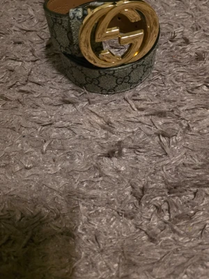 Guld beige bälte - Snyggtbälte med klassiskt -mönster och stort guldfärgat Guld-spänne. Bältet är i beige och grå toner med tydligt tryckt logomönster. Perfekt accessoar för att lyfta din outfit.