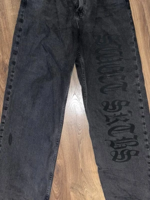 Svarta jeans från Sweet SKTBS - Säljer ett par svarta jeans från Sweet SKTBS med stor tryckt logga längs ena benet. Jeansen har en loose fit och är tillverkade i kraftigt denimtyg. Perfekta för dig som gillar streetwear och vill sticka ut med en unik detalj.