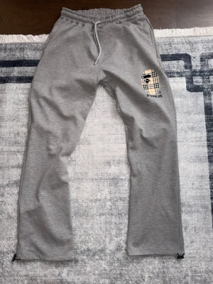 Grå mjukisbyxor med rutigt tryck - Säljer ett par grå mjukisbyxor med elastisk midja och dragsko. På vänster lår finns ett rutigt tryck med en apa och texten 'BY MONKEY APE'. Byxorna har raka ben och en bakficka. Perfekta för chill dagar eller streetwear-stil.