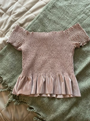 Ljusrosa offshoulder topp med volang - Supersöt ljusrosa offshoulder topp med smock över hela kroppen och volang nertill. Toppen har korta ärmar och är perfekt för sommarens alla tillfällen. Materialet känns mjukt och stretchigt, troligen polyester eller bomull.
