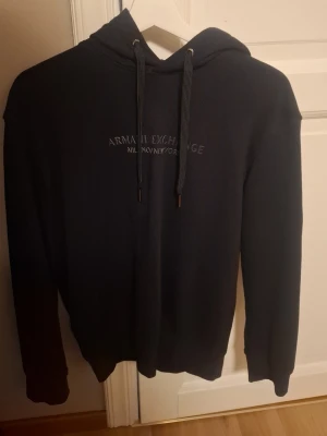 Svart hoodie från Armani Exchange - Snygg svart hoodie från Armani Exchange med broderad text 'Armani Exchange Milano/New York' på bröstet. Klassisk modell med huva och dragsnören, samt ribbade muddar vid ärmar och nederkant. Perfekt för en stilren och avslappnad look. Använt ett par gånger gillade inte den så säljer. Var jävligt dyr när jag köpte den men minns inte exakt, men duger inte priset så lägg ett bud