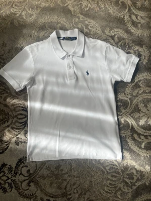 Vit pikétröja från Polo Ralph Lauren - Klassisk vit pikétröja från Polo Ralph Lauren med korta ärmar och krage. Tröjan har knäppning med två knappar framtill och den ikoniska lilla logotypen broderad på bröstet. Perfekt till jeans eller shorts för en clean look.