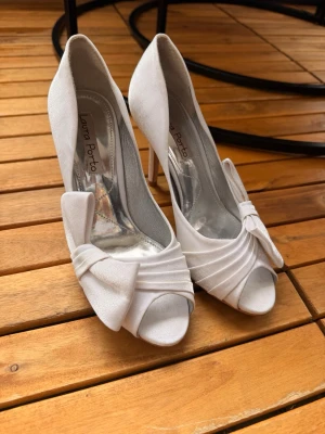 Vita peep-toe pumps med rosett - Säljer ett par eleganta vita pumps från Laura Porto med peep-toe och hög smal klack. Skorna har en stor dekorativ rosett framtill och fina draperade detaljer i satinliknande material. Perfekta för dig som vill ha en chic och stilren look.
