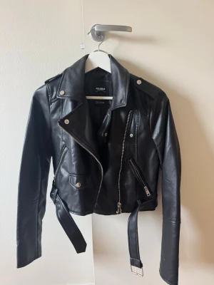 Svart skinnjacka från Pull&Bear - Snygg svart skinnjacka från Pull&Bear med klassisk bikerstil, silvriga dragkedjor och knappar samt bälte i midjan. Jackan har axelklaffar och två snedställda fickor med dragkedja. 