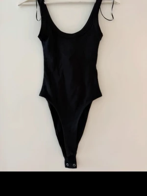 Svart basic body med smala axelband - En enkel och stilren svart body med smala axelband och rundad urringning. Bodyn har hög skärning vid benen och knäppning med tryckknappar i grenen. Perfekt att matcha med jeans eller kjol för en clean look.