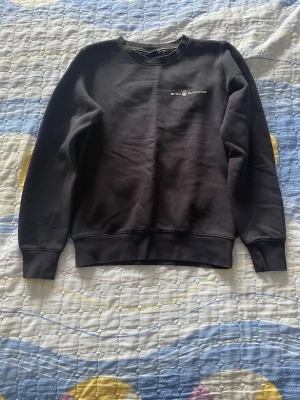 Svart Sail Racing stickad tröja - Svart stickad tröja från Sail Racing med rund halsringning och diskret logga på bröstet. Tröjan har långa ärmar och ribbade muddar vid ärmslut och nederkant. Perfekt för en stilren och avslappnad look.