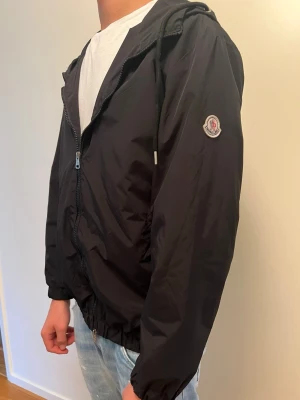 Svart vindjacka med huva Moncler - Snygg svart vindjacka från Moncler med huva och dragsko. Jackan har dragkedja framtill, elastiska muddar och Moncler-logga på ärmen. På huvan finns en röd, vit och blå rand som detalj. Tillverkad i lätt, vindtätt material.