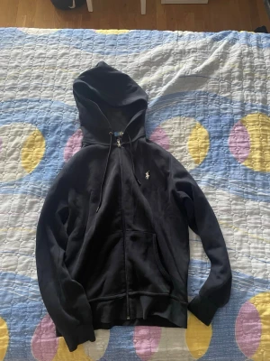 Polo Ralph Lauren Zip Up - Svart hoodie med dragkedja framtill och fickor på magen. Klassisk design med huva och justerbara snören. Liten vit logga broderad på bröstet. Tillverkad i mjukt material som känns skönt mot huden. Perfekt för en avslappnad och stilren look.