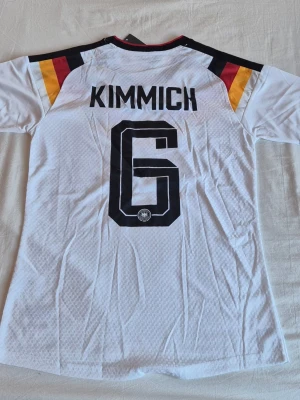 Tysklands matchtröja Kimmich nr 6 - Säljer en vit Tyskland matchtröja från Adidas med Kimmich och nummer 6 på ryggen. Tröjan har svarta, röda och gula detaljer på axlarna, samt Adidas-logga på bröstet. Tillverkad i lätt och ventilerande material, perfekt för fotboll eller träning.