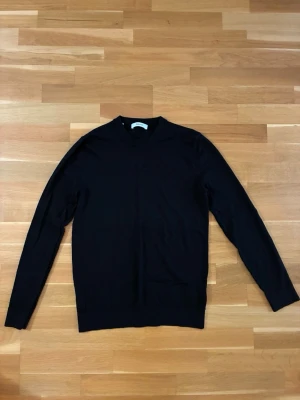 Svart stickad tröja - En stilren svart stickad tröja med rund hals och långa ärmar. tillverkad i merinoull. Tröjan har ribbade muddar vid ärmslut och nederkant, vilket ger en klassisk och enkel look. Perfekt att matcha med jeans.
