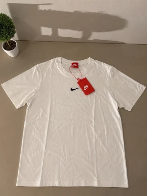 Vit Nike t-shirt med svart logo - Fräsch vit t-shirt från Nike med klassisk svart Swoosh-logga broderad på bröstet. T-shirten har rund halsringning och korta ärmar. Tillverkad i mjuk bomull för skön känsla och enkel stil.