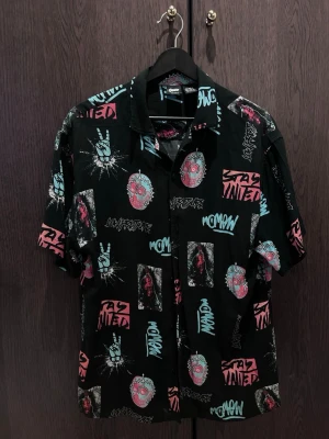 Svart skjorta med grafiskt tryck Pull&Bear - Cool svart skjorta från Pull&Bear med färgglatt grafiskt tryck i turkos, rosa och vitt. Skjortan har korta ärmar, klassisk krage och knäppning framtill. Perfekt för dig som gillar streetwear och unika prints.