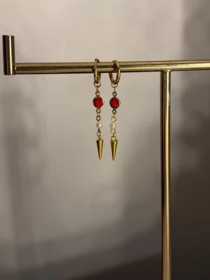 Guldörhängen med röda stenar och pärlor - Snygga örhängen i guldfärgad metall med runda röda stenar, små vita pärlor och spetsiga hängen. Perfekta för dig som vill ha något unikt och trendigt. Örhängena har en cool kombination av färger och former som sticker ut.