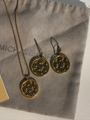 Michael Kors halsband med örhängen - Snyggt halsband + örhängen från Michael Kors i guldfärgad metall med ett runt hänge. Hänget har ett detaljerat geometriskt mönster med små pärlliknande detaljer i mitten av varje sektion. Perfekt för dig som gillar stilrena och moderna accessoarer.